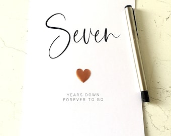 May include: A white card with the word "Seven" in elegant black script, a copper heart, and the text "YEARS DOWN FOREVER TO GO." A silver pen rests on the card.