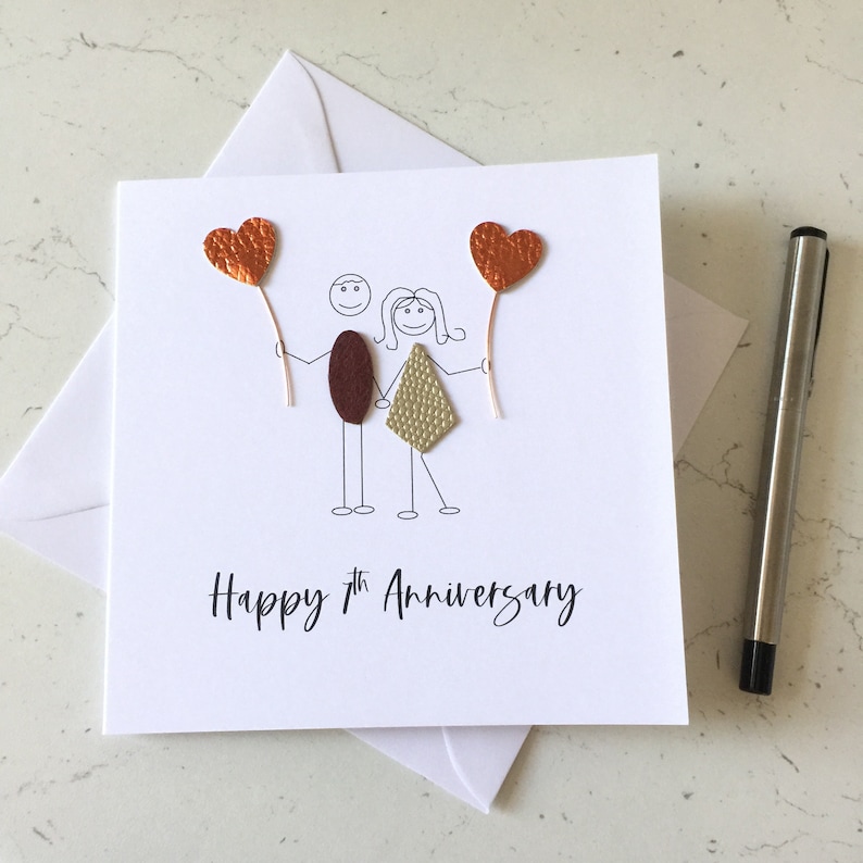 May include: A white card with a hand-drawn illustration of a couple holding red heart balloons. The text "Happy 7th Anniversary" is written below the illustration.