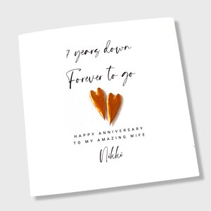 May include: A white square greeting card featuring two metallic copper-colored heart shapes in the center. The text reads: 7 years down, Forever to go, HAPPY ANNIVERSARY TO MY AMAZING WIFE, and the name Nikki in a cursive script font.