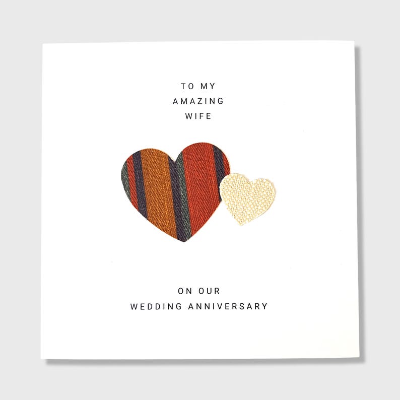 Pu&ograve; includere: Un biglietto bianco con il testo "To my amazing wife on our wedding anniversary." Due cuori sono sul biglietto, uno &egrave; di tessuto a righe rosso, arancione e nero e l'altro &egrave; un cuore bianco.