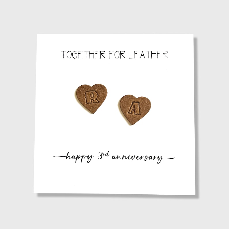 May include: A white card with the text "TOGETHER FOR LEATHER" and "happy 3rd anniversary". Two brown heart-shaped leather pieces with the letters "R" and "A" are on the card. The card is on a white background.