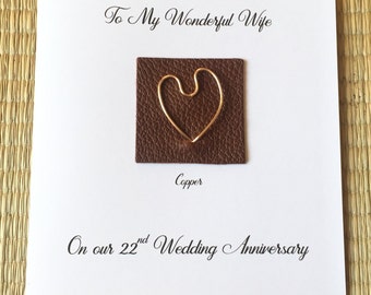 May include: A white card with the text "To My Wonderful Wife" and "On our 22nd Wedding Anniversary". A small brown leather square with a gold wire heart shape is attached to the card. The text "Copper" is written below the leather square.