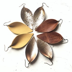 May include: Six pairs of leather earrings in various colors, including gold, brown, and yellow. The earrings are shaped like leaves and have a simple, elegant design.