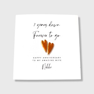 May include: A white square greeting card featuring the black text 7 years down Forever to go, two metallic copper-colored heart shapes in the center, and the text HAPPY ANNIVERSARY TO MY AMAZING WIFE followed by the name Nikki.