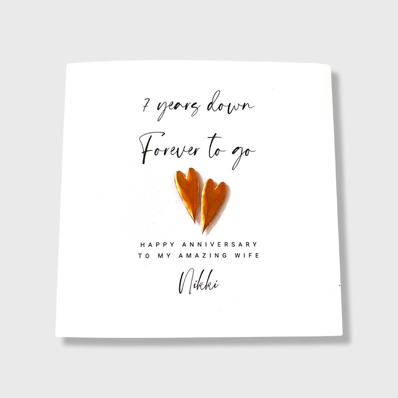 May include: A white square greeting card featuring the black text 7 years down Forever to go, two metallic copper-colored heart shapes in the center, and the text HAPPY ANNIVERSARY TO MY AMAZING WIFE followed by the name Nikki.