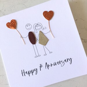 May include: A white card with a hand-drawn illustration of a couple holding hands. The couple is made of brown and gold felt. The card has two copper hearts with copper wire stems. The text "Happy 7th Anniversary" is written in black ink.