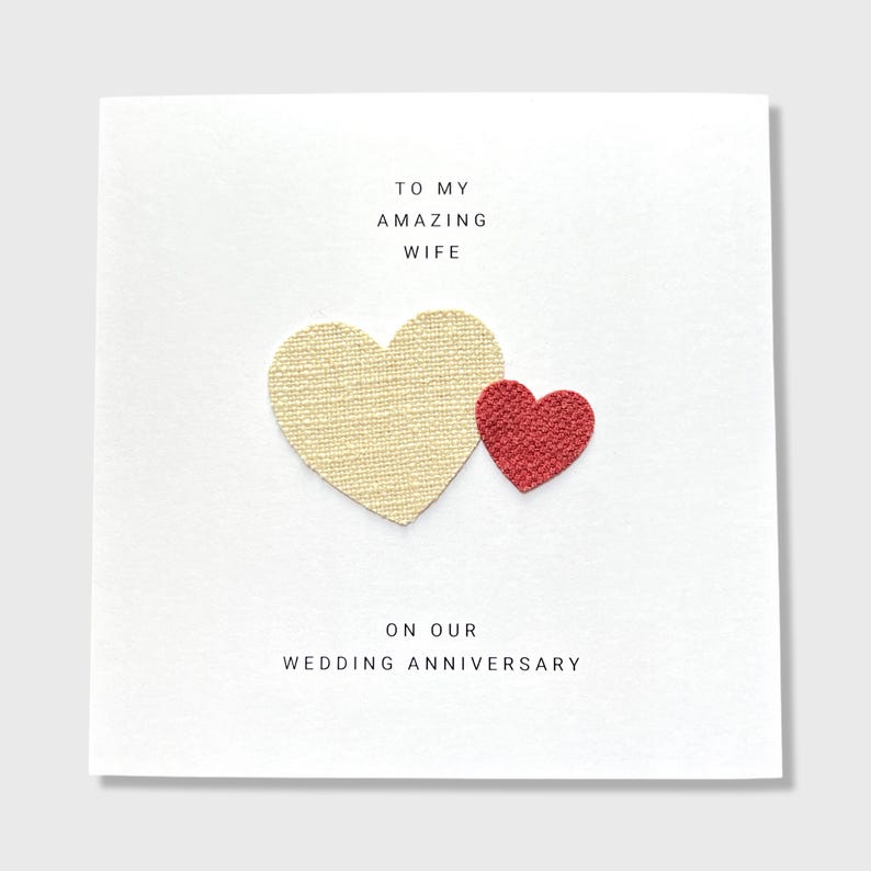 Pu&ograve; includere: Un biglietto bianco con il testo "To my amazing wife on our wedding anniversary." Due cuori sono attaccati al biglietto, un cuore grande color crema e un cuore rosso pi&ugrave; piccolo.