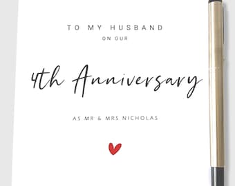 May include: A white greeting card with the text "TO MY HUSBAND ON OUR 4th ANNIVERSARY" in black script. Below, it reads "AS MR & MRS NICHOLAS" with a small red heart. A gold pen rests on the right side.