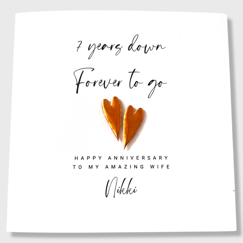 May include: A white greeting card featuring the black text 7 years down Forever to go, two metallic copper-colored heart shapes, and the black text HAPPY ANNIVERSARY TO MY AMAZING WIFE followed by the name Nikki in a script font.