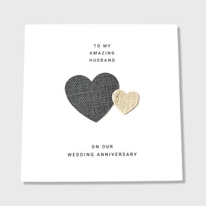 Pu&ograve; includere: Un biglietto bianco con il testo "To my amazing husband on our wedding anniversary." Due cuori di stoffa sono attaccati al biglietto, uno grigio e uno beige.
