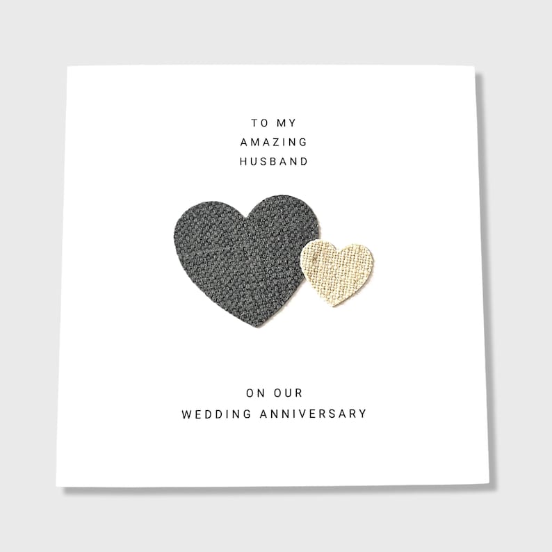 Pu&ograve; includere: Un biglietto bianco con il testo "To my amazing husband on our wedding anniversary." Due cuori di stoffa sono attaccati al biglietto, uno grigio e uno beige.