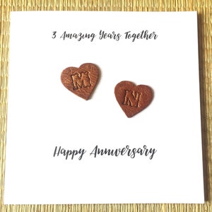 May include: A white square card featuring the text 3 Amazing Years Together at the top and Happy Anniversary at the bottom. Two brown leather heart-shaped cutouts are attached to the center, each embossed with a single letter, M and N.