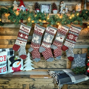 Peut inclure: Cinq chaussettes de Noël personnalisées avec des motifs en tricot rouge, blanc et gris. Les chaussettes sont accrochées à un mur en bois avec une guirlande de lumières. Les chaussettes sont étiquetées "Mama", "Ryan", "Fynn", "Jamie" et "Mommy".