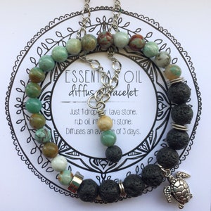 Könnte beinhalten: Zwei Armbänder, eines mit grünen und braunen Steinen und das andere mit schwarzen Lavasteinen. Das Lavasteinarmband hat einen silbernen Schildkrötenanhänger. Die Armbänder befinden sich auf einer weißen Karte mit dem Text "ESSENTIAL OIL diffusracelet" und Gebrauchsanweisungen.