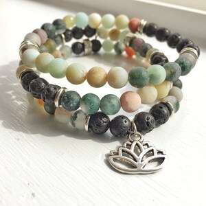 Op de afbeelding: Een drievoudig armband met groene, bruine en witte kralen, afgewisseld met zwarte lava kralen. Een zilveren lotusbloem bedel hangt aan de onderkant van de armband.
