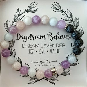 May include: A lavender, pink and white gemstone bracelet with black lava beads and a silver charm that says "Daydream Believer" and "Dream Lavender Joy Love Healing". The bracelet is on a white background with a floral design.