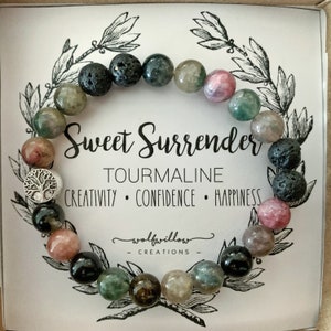 May include: A bracelet made with green, black, and pink tourmaline beads and black lava rock beads. The bracelet has a silver charm in the shape of a tree of life. The card reads "Sweet Surrender Tourmaline Creativity Confidence Happiness wolfwillow Creations".
