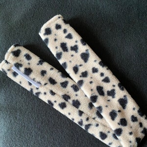 May include: Two beige and black fuzzy fabric arm warmers with a leopard print pattern. The arm warmers are folded in half and the text "The Shughes" is visible on one of the arm warmers.