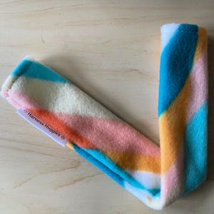 May include: A colorful, soft, fleece harness accessory with a striped pattern in shades of blue, orange, pink, and white. The product has a tag that reads "Harness Huggies".