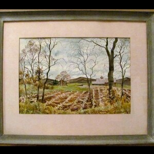 May include: A framed watercolor painting of a rural landscape. The artwork depicts a field with rows of harvested crops, bare trees, and a building in the distance. The frame is a muted green color.