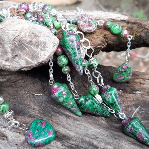 May include: A silver chain necklace with green and red gemstone beads and pendants. The necklace features a variety of shapes, including hearts, teardrops, and points. The gemstones are a mix of green and red, with some having a speckled appearance.