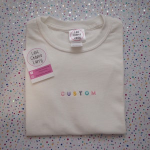 May include: Cream-coloured t-shirt with the word "CUSTOM" embroidered in rainbow colours. The shirt has a crew neck and is folded neatly. A tag with the text "Last Chance Larry" is attached. A small card with the same logo is also visible.