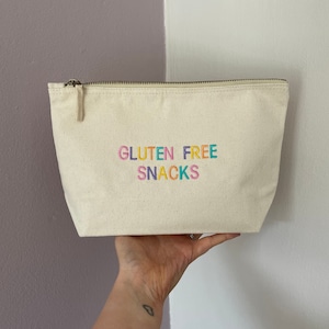 May include: A cream-coloured canvas pouch with a zip closure. The pouch features the embroidered text "GLUTEN FREE SNACKS" in rainbow colours. The background is a split wall, half purple and half white.