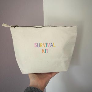 May include: An off-white canvas zipped pouch featuring the embroidered text SURVIVAL KIT in pastel pink, blue, green, and yellow thread. The pouch has a dark metal zip closure at the top and a simple, structured trapezoidal shape.