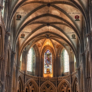 May include: A high vaulted ceiling with intricate stonework and stained glass windows in a cathedral. The ceiling is made of arched stone and the windows are tall and narrow. The light from the windows illuminates the interior of the cathedral.