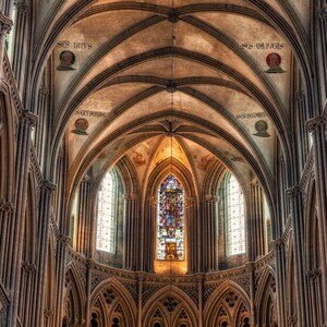 May include: A high vaulted ceiling with intricate stonework and stained glass windows in a cathedral. The ceiling is made of arched stone and the windows are tall and narrow. The light from the windows illuminates the interior of the cathedral.