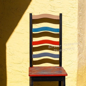 May include: A black wooden chair with a red, blue, and brown painted back and seat. The chair is sitting in front of a yellow wall.