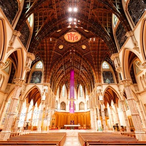 May include: An interior shot of a cathedral with a high, detailed wooden ceiling. The ceiling is carved with a repeating pattern. Stained-glass windows allow sunlight to enter. The church has rows of wooden pews and a central aisle leading to the altar.