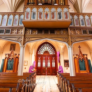 May include: A large wooden pipe organ with gold pipes is situated above the entrance to a church. The church has a vaulted ceiling and wooden pews. The entrance to the church is framed by two large wooden doors with stained glass windows.