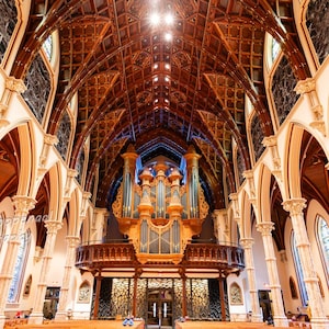 May include: An interior shot of a cathedral, showcasing a high, detailed wooden ceiling. The space includes tall, arched columns and stained-glass windows. A large pipe organ is centrally positioned above a balcony, with decorative elements and warm lighting.