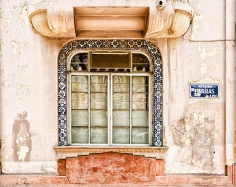 May include: An exterior shot of a building with a window. The window is framed with blue and white patterned tiles. The building's facade is painted in shades of pink and peach, with a street sign that reads "AVENIDA PARRAS".