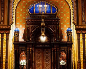 Może przedstawiać: Widok w dół alei synagogi z ozdobnymi złotymi i niebieskimi dekoracjami. Aleja jest wyłożona drewnianymi ławkami i prowadzi do dużej drewnianej arki z wiszącym nad nią złotym świecznikiem.