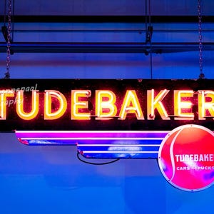 Könnte beinhalten: Ein Vintage-Neon-Schild mit dem Wort "Studebaker" in roten Neonbuchstaben. Das Schild hängt an einer Kette und hat ein rot-weißes Logo mit dem Text "Studebaker Cars Trucks" darunter.