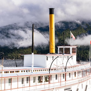 Può includere: Un battello a vapore bianco con un camino giallo e nero, attraccato a un molo. La barca ha più ponti e finestre. La bandiera canadese sventola dall'albero maestro. La barca è di fronte a una catena montuosa con le nuvole.