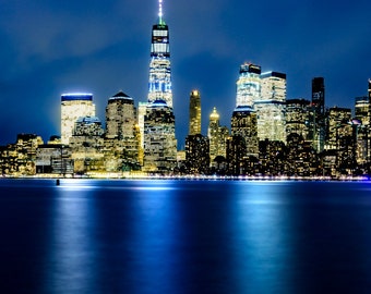 Pode incluir: Uma vista noturna do horizonte da cidade de Nova York com a Freedom Tower iluminada no centro. A água em primeiro plano reflete as luzes da cidade.