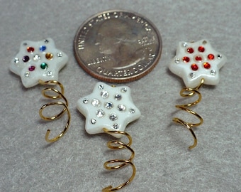 Op de afbeelding: Drie stervormige haaraccessoires met witte centra en juwelen accenten. Eén heeft kleurrijke juwelen, één heeft heldere juwelen en één heeft rode juwelen. Elke ster is bevestigd aan een gouden spiraaldraad.