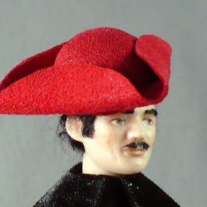 May include: A miniature doll with a red hat and a black coat. The hat is a vibrant red with a textured surface and a wide brim. The doll has a mustache and dark hair. The coat is black and appears to be made of velvet.
