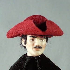 May include: A red felt tricorn hat with a wide brim and a deep crown. The hat is worn by a person with dark hair and a mustache.