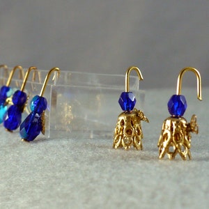 Op de afbeelding: Goudkleurige oorbellen met blauwe kristallen kralen. De oorbellen hebben een decoratieve goudkleurige belvormige basis. De oorbellen worden gepresenteerd op een lichtgrijze ondergrond.