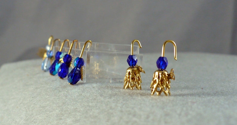 Op de afbeelding: Goudkleurige oorbellen met blauwe kristallen kralen. De oorbellen hebben een decoratieve goudkleurige belvormige basis. De oorbellen worden gepresenteerd op een lichtgrijze ondergrond.