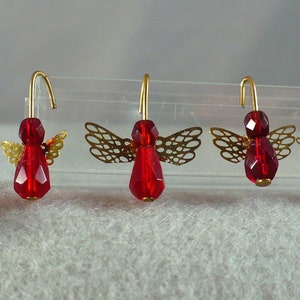 May include: Five red beaded angel earrings with gold-colored wire hooks and filigree wings. The angels have faceted red bodies and small gold wings. The earrings are displayed on a clear plastic stand against a soft, light background.