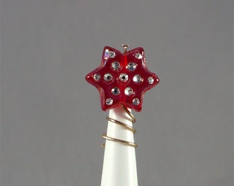Op de afbeelding: Een rode, stervormige kerstboomtop met heldere, sprankelende accenten. De ster is gemonteerd op een witte kegel met gouddraadwikkeling. Een kleine metalen pin bevestigt de ster aan de kegel.
