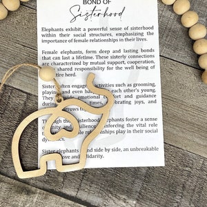 May include: A wooden cutout of two elephants side by side, with the text "The Unbreakable Bond of Sisterhood" and a description of the bond between female elephants. The cutout is on a white background with a string of wooden beads.