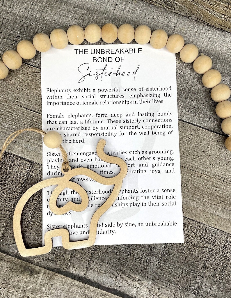 May include: A wooden cutout of two elephants side by side, with the text "The Unbreakable Bond of Sisterhood" and a description of the bond between female elephants. The cutout is on a white background with a string of wooden beads.