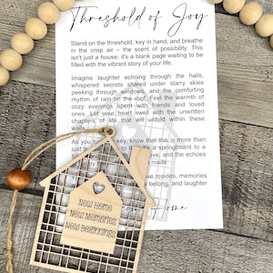 May include: A wooden house-shaped ornament with the text "New Home, New Memories, New Beginnings" on it. The ornament is hanging from a string and has a white card attached to it with the text "Threshold of Joy" and a poem about the joy of a new home.