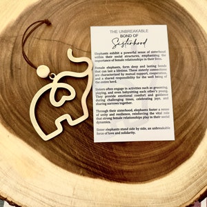 May include: A wooden elephant ornament with a heart-shaped cutout hangs from a brown cord. The ornament is on a wooden surface. The text on the card reads: "The Unbreakable Bond of Sisterhood. Elephants exhibit a powerful sense of sisterhood within their social structures, emphasizing the importance of female relationships in their lives."